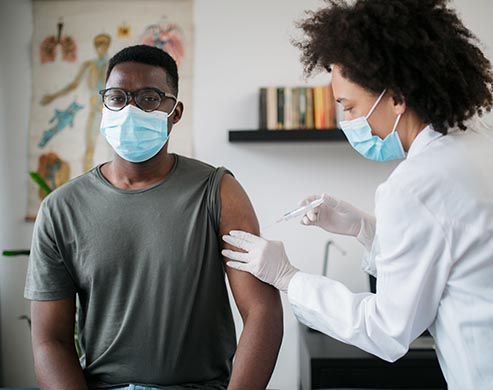 Portrait of a young African American male patient sitting in a medical clinic and is being given the Covid 19 vaccine in his shoulder by a female African American doctor, both wearing protective face masks